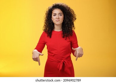 Portrait of gloomy girl with sad face pursing lips down frowning disappointed and pointing down with regret and unsatisfied expression, missing opportunity feeling upset over yellow background