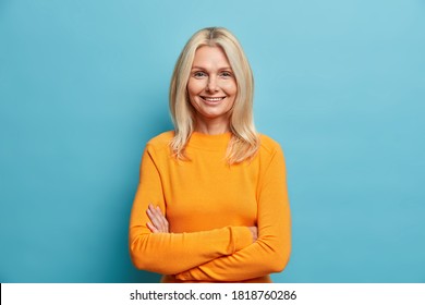 Portrait Of Good Looking Blonde Woman Stands Self Assured Against Blue Studio Background Smiles Gently And Keeps Arms Folded Glad To Meet Old Friend. People Age Beauty Sincere Emotions Concept