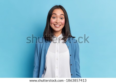 Portrait of good looking brunette woman smiles toothily hears something pleasant wears casual jumper and shirt models indoor against blue background. Feminine girl has upbeat mood. Positive emotions