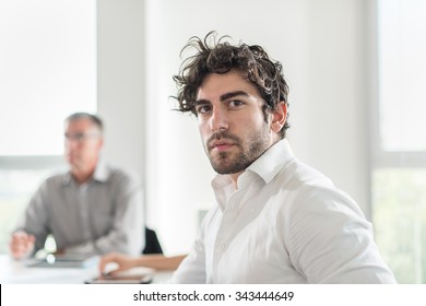 Portrait of a good looking business man with dark curly hair and beard. He is turned back on his chair looking at camera in a luminous white office. Coworkers are sitting at the table behind him