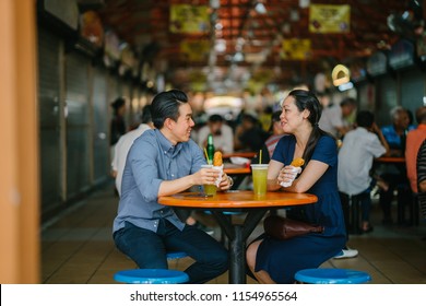 Portrait of a good looking Chinese Asian couple enjoying a snack at a hawker center during the weekend in Singapore, Asia. They are smiling as they talk and enjoy sugarcane juice and fried bananas. 