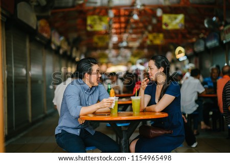 Portrait of a good looking Chinese Asian couple enjoying a snack at a hawker center during the weekend in Singapore, Asia. They are smiling as they talk and enjoy sugarcane juice and fried bananas. 