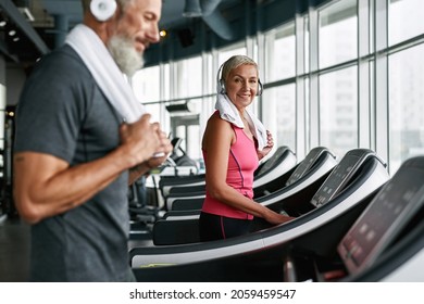 Portrait Of Good Looking And Fit Elder Woman Working Out On Treadmill. Stylish Senior Woman Looking Younger Than Her Age. Man With Towel On Shoulder On Foreground, Blured. Couple In Gym.