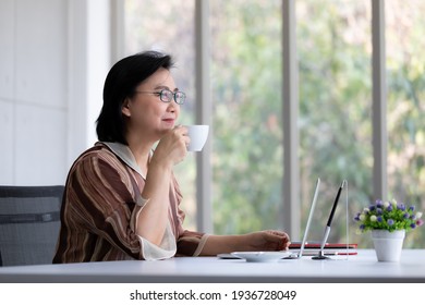 Portrait Of Good Looking And Kindly Warm Feeling Asian Senior Woman Wearing Eyeglasses Sitting And Drinking Coffee At Working Desk.