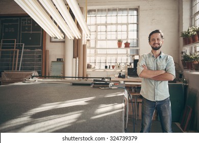 Portrait of a good looking male designer standing in his workshop studio with his arms folded and smiling confidently at the camera