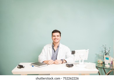 Portrait Of A Good Looking Male Hispanic Therapist In A Lab Coat Sitting At A Front Desk In A Health Spa