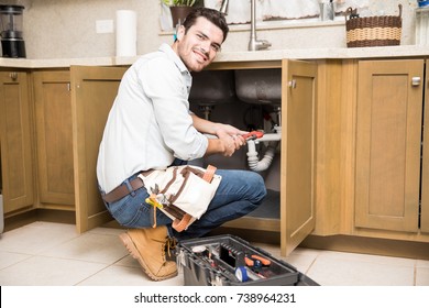 Portrait Of A Good Looking Male Plumber Fixing A Leaky Sink In A Kitchen And Smiling