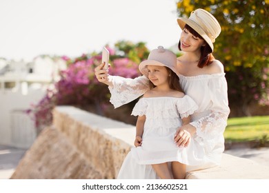 Portrait Of Good Looking Mother Taking Photo With Daughter On Mobile Phone While Sitting In The Tropical Park During Summer Vacation. Happy Mom With Daughter Use Mobile Phone While Sitting On Stone.