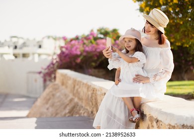 Portrait Of Good Looking Mother Taking Photo With Daughter On Mobile Phone While Sitting In The Tropical Park During Summer Vacation. Happy Mom With Daughter Use Mobile Phone While Sitting On Stone.