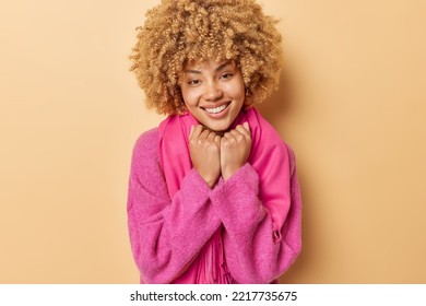 Portrait Of Good Looking Woman With Curly Hair Keeps Hands Under Chin Smiles Toothily Wears Pink Jumper And Scarf Isolated Over Beige Background. Positive Emotions And Genuine Happiness Concept