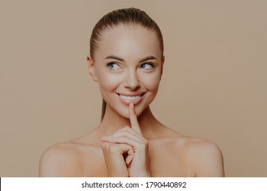 Portrait Of Good Looking Woman Looks Aside Mysteriously, Keeps Finger On Chin And Smiles Broadly, Looks Away And Shares Secret Of Beauty, Has Perfect Glowing Skin, Naked Body, Natural Makeup