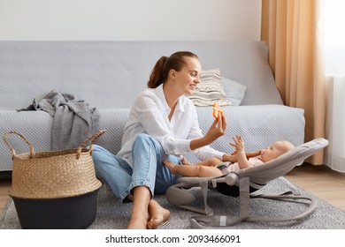 Portrait of good looking woman wearing white shirt and jeans sitting on floor near cough and playing with her infant baby, showing toy to her daughter in rocking chair. - Shutterstock ID 2093466091