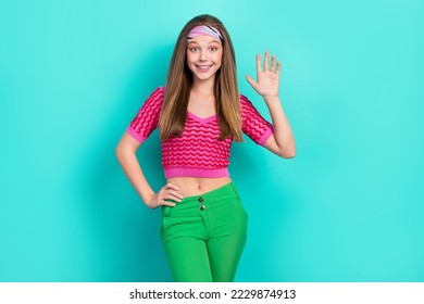 Portrait of good mood positive girl with long hairstyle dressed pink top headband wave palm arm on waist isolated on teal color background