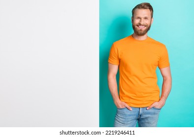 Portrait Of Good Mood Satisfied Guy With Blond Hairdo Wear Orange T-shirt Hands In Pockets Placard Isolated On Teal Color Background