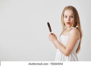 Portrait of good-looking funny small girl with blond long hair looking in camera with surprised face expression, being dirty after eating ice cream.