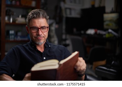 Portrait Of A Good-looking Happy Older White Man In Glasses Sitting Reading Book At Home, Indoors.