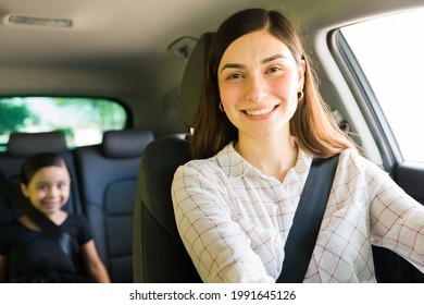 Portrait Of A Good-looking Latin Young Woman Smiling And Making Eye Contact While Driving The Car With A Little Kid In The Back