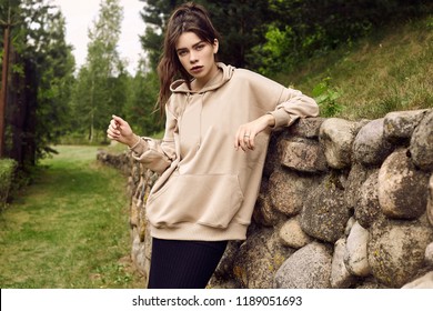 Portrait Of Gorgeous Brunette Woman In Fashion Hoodie Posing In The Garden Alley