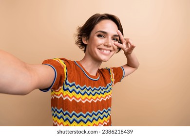 Portrait of gorgeous positive girl make selfie hand fingers demonstrate v-sign near eye isolated on beige color background