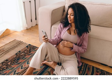 Portrait of gorgeous pregnant female with dark skin and curls sitting on carpet next to couch browsing news feed in her social media app, reading article, watching podcast for future moms - Powered by Shutterstock