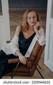 Portrait Of Gorgeous Redhead Woman In Black Evening Dress And White Shirt. Stylish Confident Mid Adult Lady Is Sitting In An Arm Chair At Home. Happy European Woman Is Going Off To A Party.