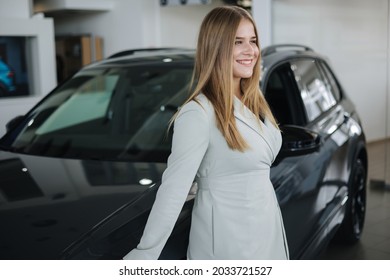 Portrait Of Gorgeous Woman In Showroom. Beautiful Salesperson Standing By Car. Blond Hair Female After Buying Car