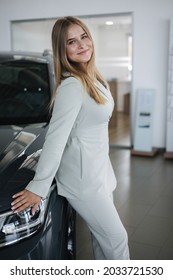 Portrait Of Gorgeous Woman In Showroom. Beautiful Salesperson Standing By Car. Blond Hair Female After Buying Car