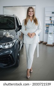 Portrait Of Gorgeous Woman In Showroom. Beautiful Salesperson Standing By Car. Blond Hair Female After Buying Car