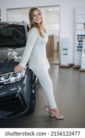 Portrait Of Gorgeous Woman In Showroom. Beautiful Salesperson Standing By Car. Blond Hair Female After Seles Car