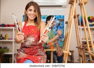 Portrait of a gorgeous young brunette wearing an apron and holding a bunch of paintbrushes in an art class