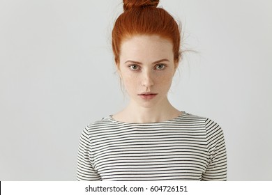 Portrait Of Gorgeous Young Female With Clean Healthy Skin With Freckles And Hair Knot Staring At Camera, Having Serious Confident Look, Dressed In Casual Striped Top. People And Lifestyle Concept