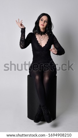 Portrait of a goth girl with dark hair wearing black dress and boots. Full length sitting pose on a studio background.