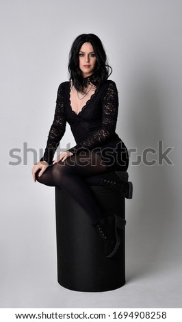 Portrait of a goth girl with dark hair wearing black dress and boots. Full length sitting pose on a studio background.