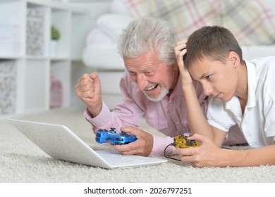 Portrait Of Grandfather And Grandson Lying On Floor And Playing Computer Games On Laptop