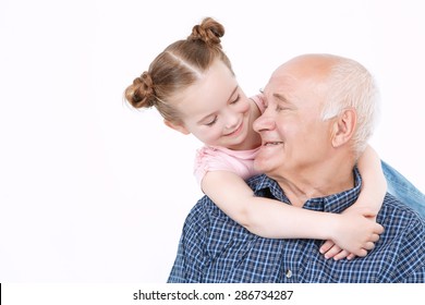 Portrait of a grandfather wearing blue checkered shirt sitting and small pretty granddaughter standing behind him smiling and hugging, looking at each other, isolated on a white background