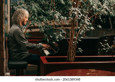 Portrait of gray-haired senior man with curly long hair is sitting at piano and playing music, enjoying pleasant evening at hotel, side view. Lifestyle photo