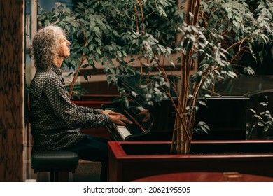  Portrait of gray-haired senior man with curly long hair is sitting at piano and playing music, enjoying pleasant evening at hotel. Lifestyle photo