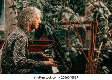  Portrait of gray-haired senior man with curly long hair is sitting at piano and playing music, enjoying pleasant evening at hotel, side view. Lifestyle photo
