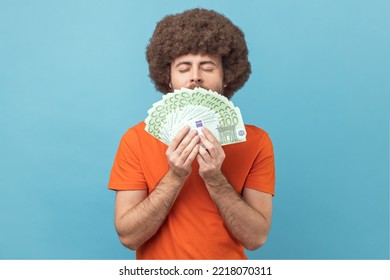 Portrait Of Greedy Man With Afro Hairstyle In Orange T-shirt Smelling Euro Banknotes With Pleasure Expression, Enjoying Big Money Profit And Rich Life. Indoor Studio Shot Isolated On Blue Background.