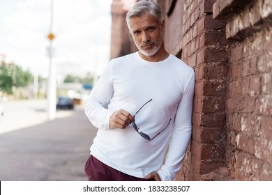 Portrait Of A Grey-haired Mature Handsome Man In Jeans And White T-shirt Leaning To The Old Wall