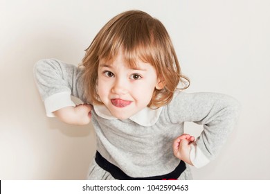 Portrait Of Grimacing Little Baby Girl Shows Tongue On Light Background