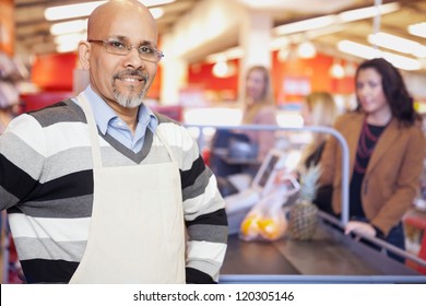 Portrait Of A Grocery Store Cashier Standing At Checkout Counter With Customers In The Background