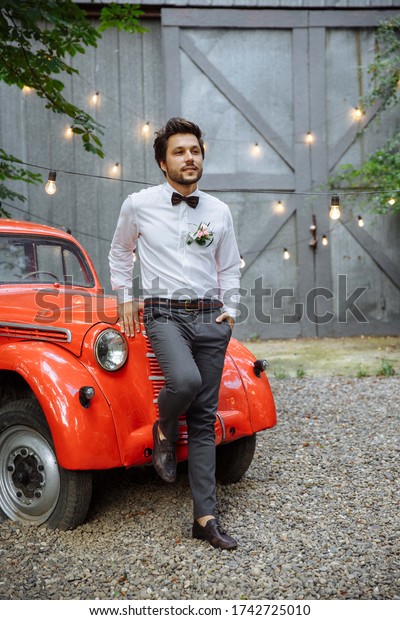 Portrait of the groom on his wedding day. In the\
background are light bulbs and a gray wall. Outdoors. Leaned on a\
red retro car
