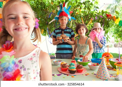 Portrait Of Group Of Children In Fancy Dresses At A Colorful Birthday Party In A Home Garden With Decorations, Holding Cake With Candle. Joyful Expressions Outdoors Lifestyle. Kids Activities And Fun.