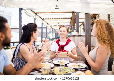 Portrait of group of friends celebrating birthday in outdoor cafe, focus on happy man holding gift box with red ribbon, copy space - Powered by Shutterstock