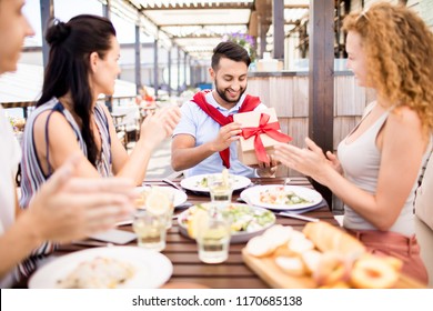 Portrait of group of friends celebrating birthday in outdoor cafe, focus on happy Middle-Eastern man opening gift box with red ribbon, copy space - Powered by Shutterstock