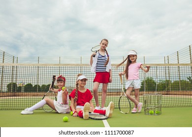 Portrait Of Group Of Girls As Tennis Players Holding Tennis Rackets Against Green Grass Of Outdoor Court. Stylish Young Teens Posing At Park. Sport Style. Teen And Kids Fashion Concept.