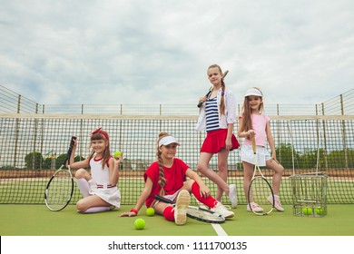 Portrait Of Group Of Girls As Tennis Players Holding Tennis Rackets Against Green Grass Of Outdoor Court. Stylish Young Teens Posing At Park. Sport Style. Teen And Kids Fashion Concept.