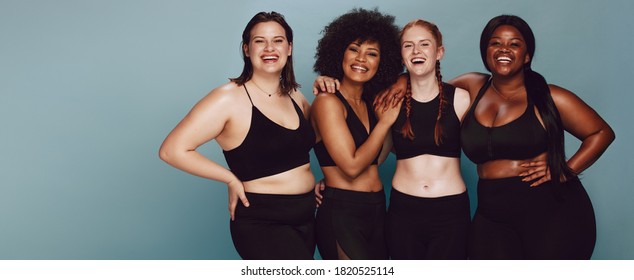 Portrait of group of women posing together in sportswear against a gray background. Multiracial females with different size standing together looking at camera and smiling.