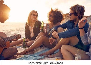 Portrait of group of young friends having a party on the beach in evening. Men and women drinking beers and listening to friend playing guitar. - Powered by Shutterstock